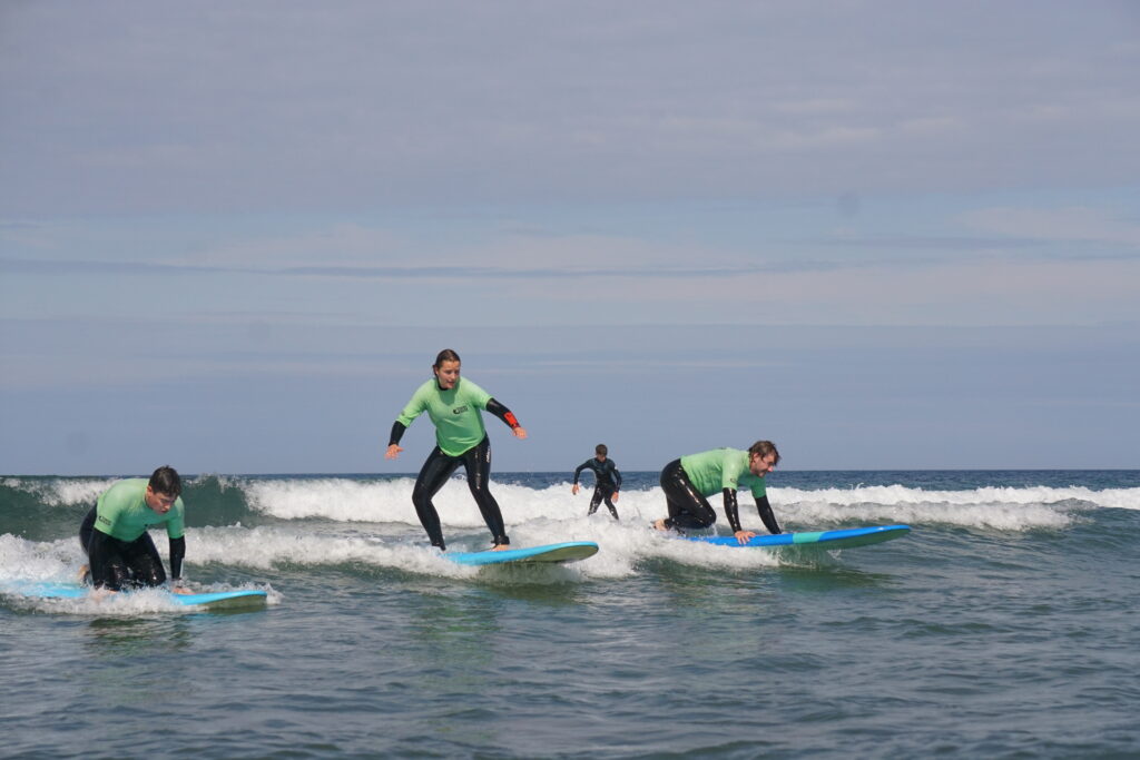 cornwall uk surfing. Three learner surfers riding a wave. One surfer is standing.