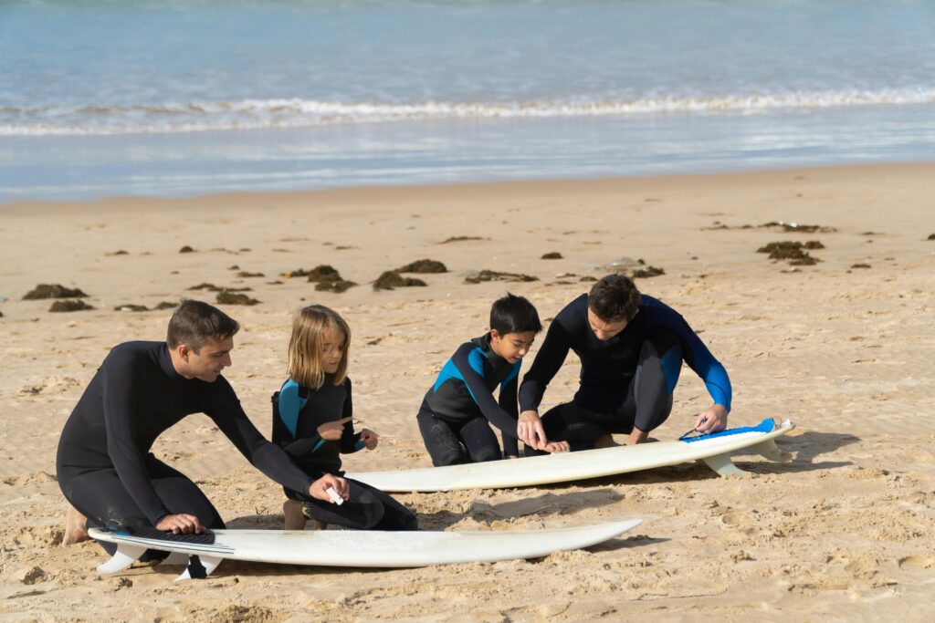 surfing lessons cornwall. Two adults show two young kids how to prepare their surfboards.