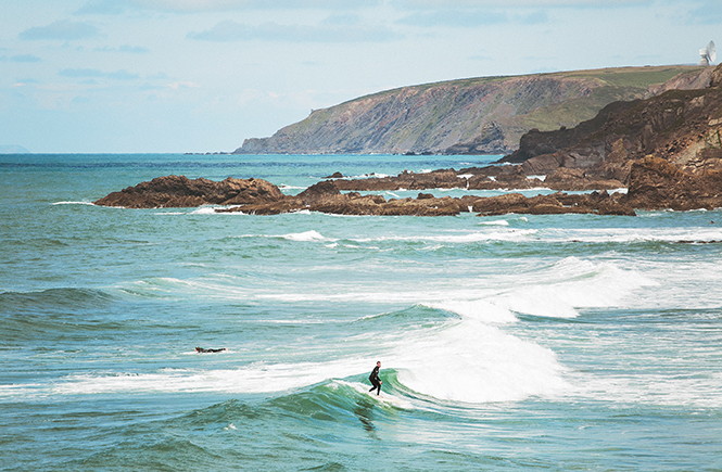 best surf beach uk. Surfer standing up and riding a waves with the clifftop coastline in the background.